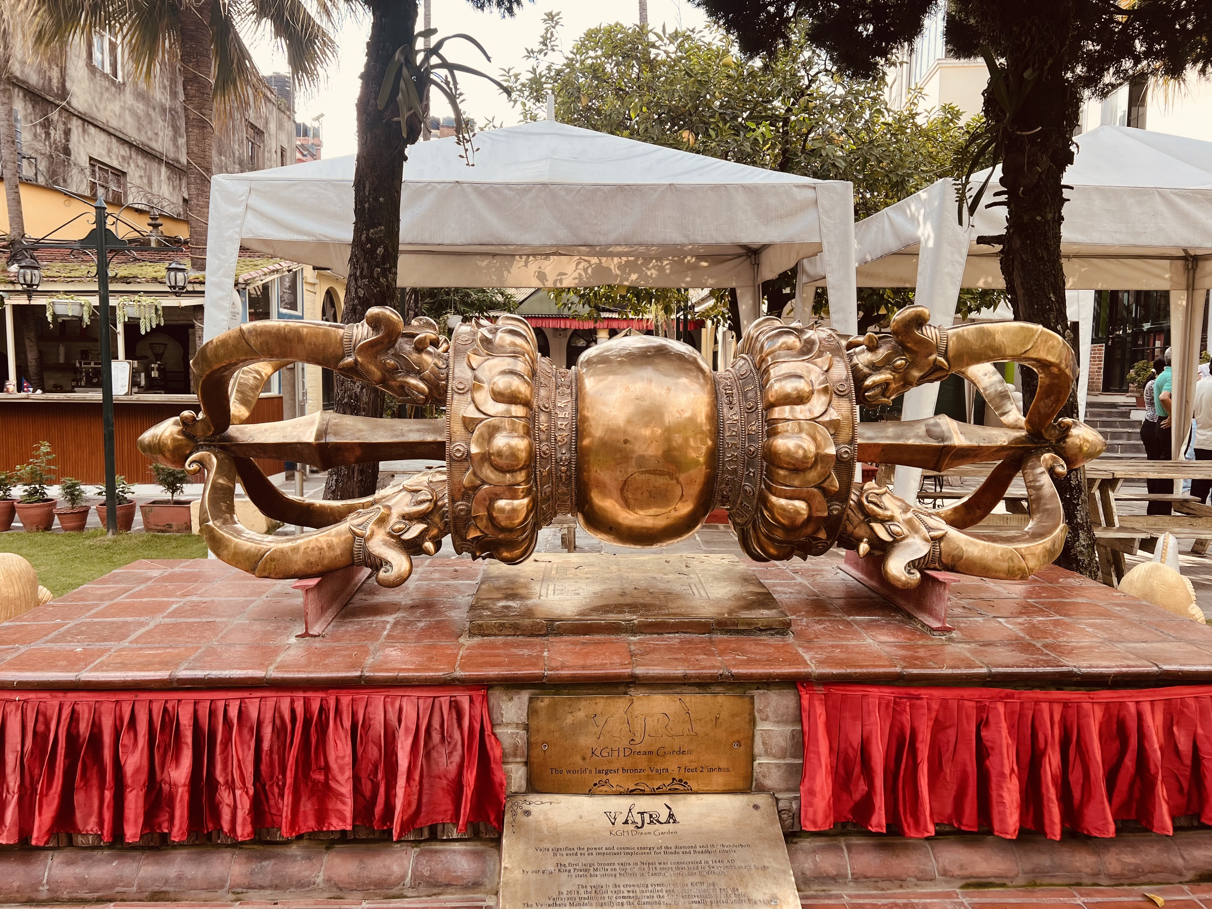 A large, ornate brass Vajra or Dorje sculpture rests on a raised, red-tiled platform with a descriptive plaque in the foreground. White tents are in the background.