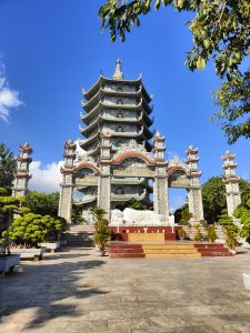 A tall multi-tiered pagoda at Chùa Linh Ứng in Da Nang, Vietnam, standing under a bright blue sky. In front of the entrance, a white reclining Buddha statue rests on a raised platform surrounded by trees and stone carvings.