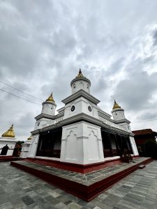 White temple with three golden domes and intricate architectural details, set against a cloudy sky. The atmosphere feels serene and timeless.