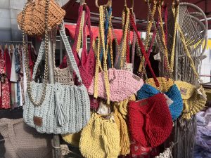 A display of many colorful, hand-crocheted handbags in various shades, including pale blue, pink, yellow, and red. They are hanging from a wooden rack, some with chunky gold chain straps.