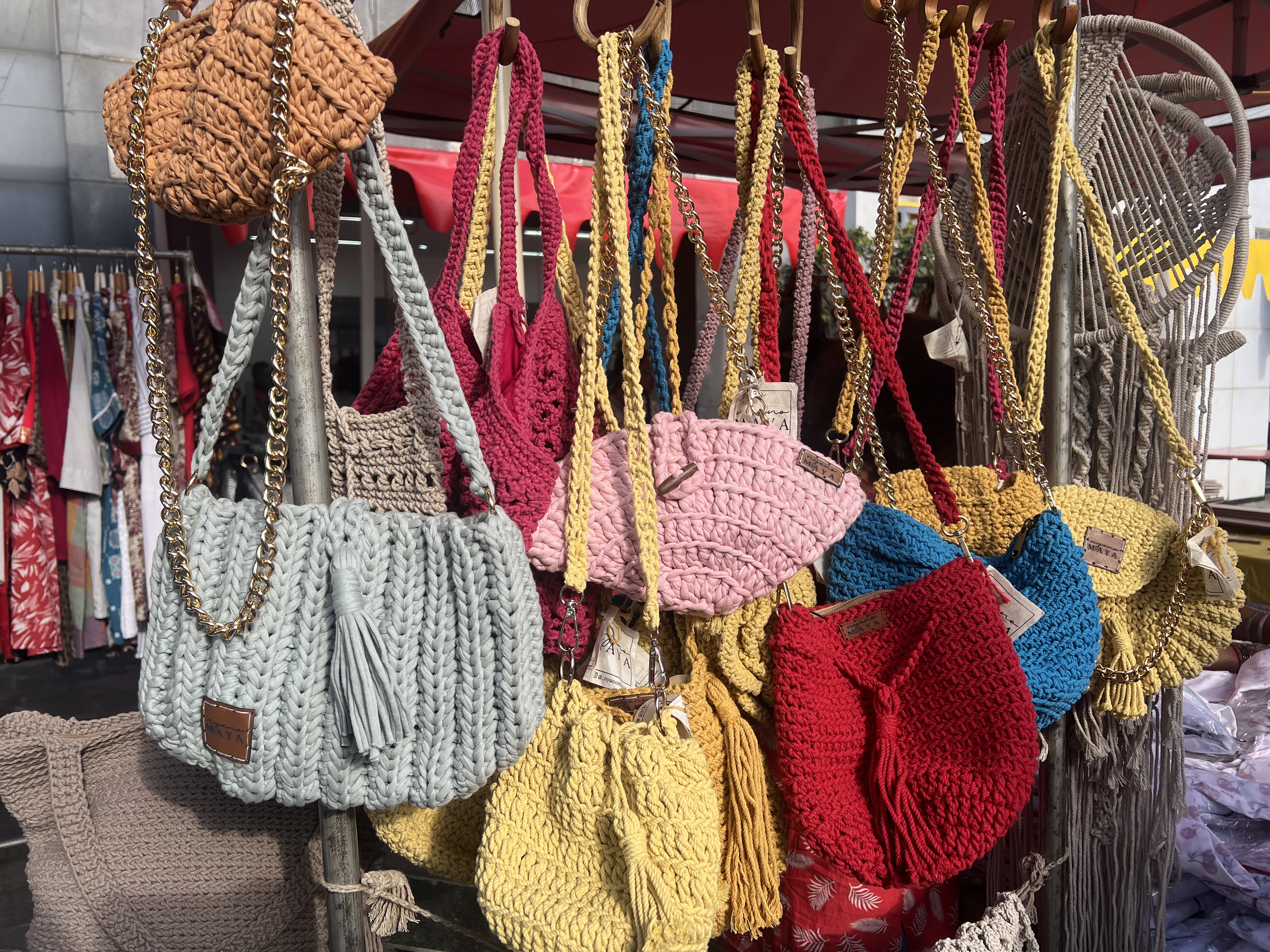 A display of many colorful, hand-crocheted handbags in various shades, including pale blue, pink, yellow, and red. They are hanging from a wooden rack, some with chunky gold chain straps.