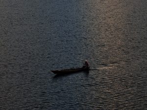 A lone boat drifts on tranquil twilight waters, where soft blue and orange hues reflect as a person calmly navigates in peaceful solitude.