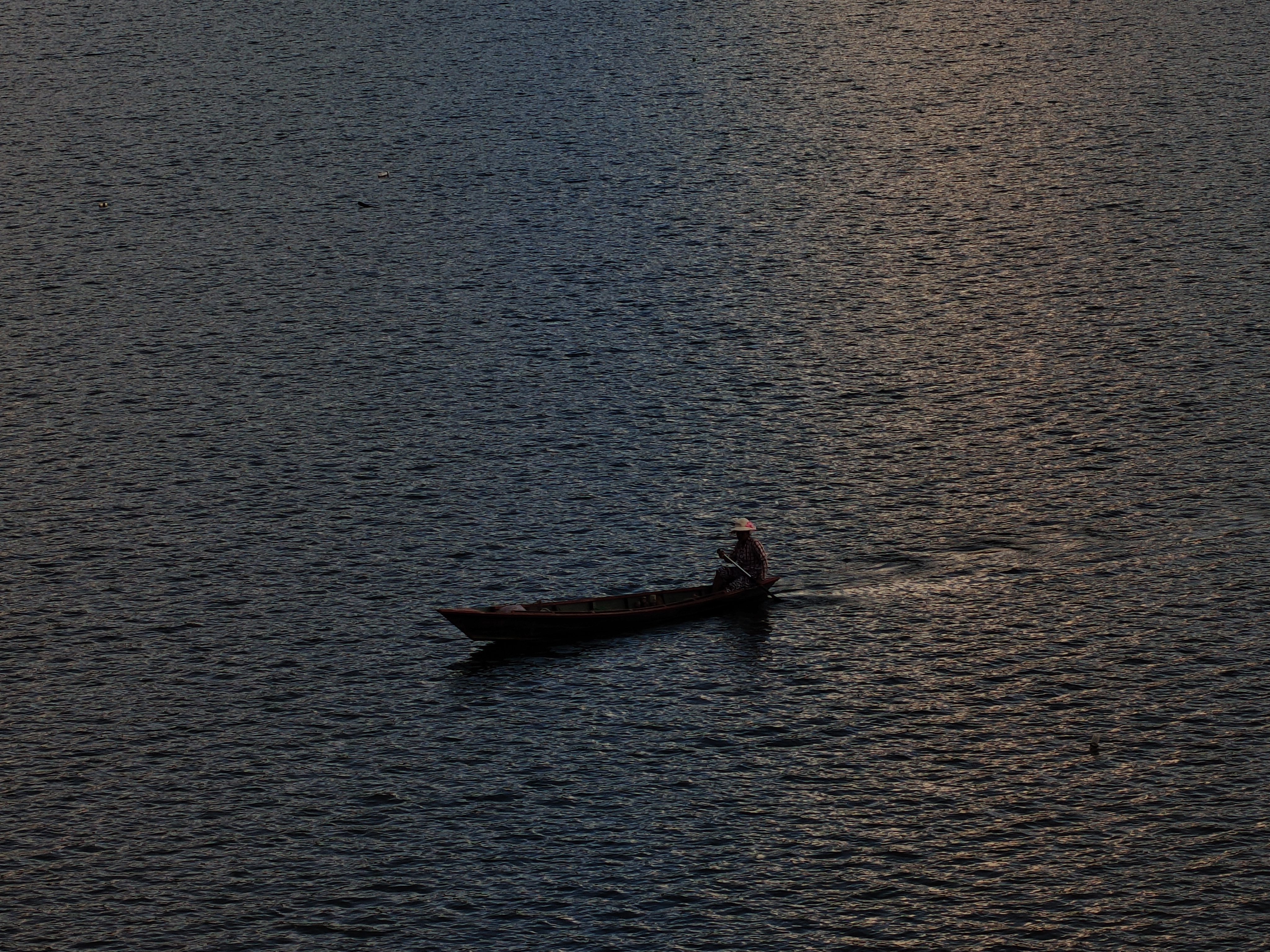 A lone boat drifts on tranquil twilight waters, where soft blue and orange hues reflect as a person calmly navigates in peaceful solitude.