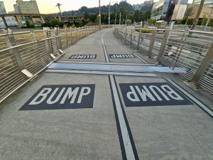 Sidewalk warning signs painted with “BUMP” in black boxes on the Tilikum Crossing Bridge in Portland. View includes fencing and city background. 