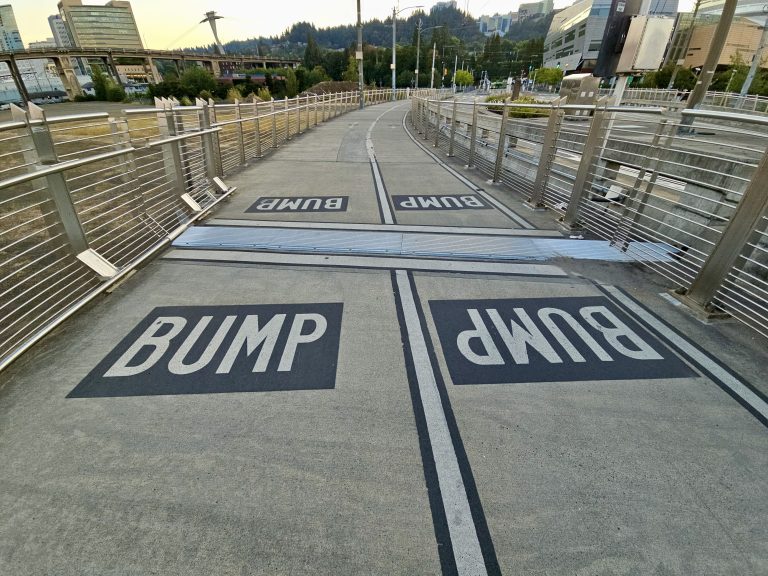 Sidewalk warning signs painted with “BUMP” in black boxes on the Tilikum Crossing Bridge in Portland. View includes fencing and city background.