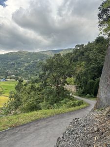 A winding road leads through lush green mountains, with patches of farmland visible in the valley below