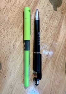 A neon green marker or highlighter and a black multi-tool pen with a stylus tip rest side-by-side on a wooden surface.
