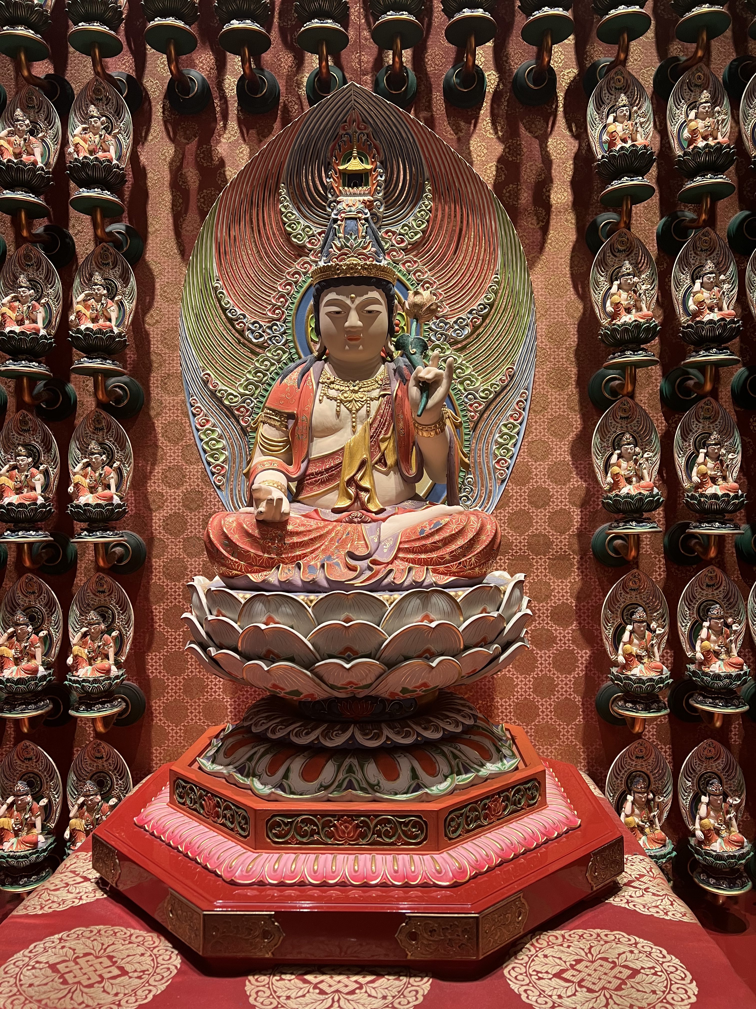 A large, brightly painted statue of a seated Buddha is centered on a lotus pedestal, surrounded by dozens of smaller, identical Buddha statues arranged in columns on a red-patterned wall.
