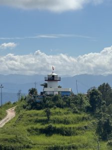 A view of a cloudy sky along with green bushy land. A small walking road leads to a tall, cemented building with a flag of Nepal fluttering at its top.