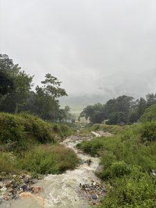 A winding stream flows through a lush green landscape, bordered by scattered rocks and vegetation. The background features misty mountains partially obscured by fog, with a field visible in the far distance. The weather appears cloudy and overcast, creating a tranquil and serene atmosphere.