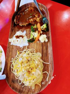 A wooden platter holds a piece of steak covered in brown sauce, a side of coleslaw, a mix of steamed vegetables, and a serving of creamy spaghetti.