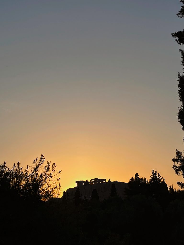 The Acropolis of Athens rises in silhouette atop the hill, framed by dark vegetation, with the sunset sky shifting from deep blue to warm orange and yellow.