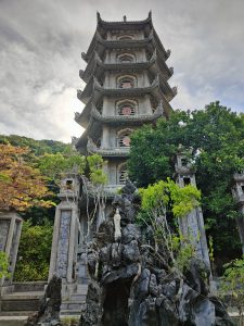 A tall stone pagoda at the Marble Mountains in Da Nang, Vietnam, surrounded by green trees. At the base, a small white Buddha statue is placed on rocky formations in front of the entrance.