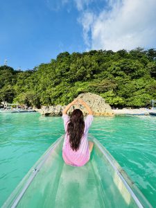 A woman with long dark hair sits in a transparent kayak on bright turquoise water, facing a lush green, rocky island under a blue sky.