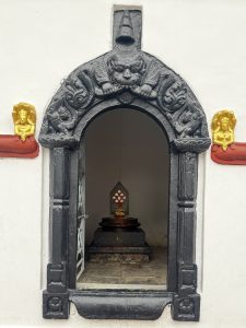 Ornate black arch with intricate carvings frames a small shrine. Inside, a golden statue on a pedestal, flanked by gold reliefs, conveys serenity and reverence.