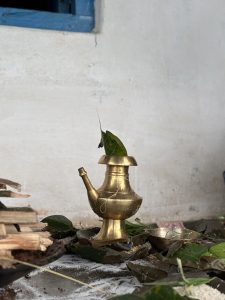 A brass water pot with a spout sits on the ground among scattered green leaves and some white powder.
