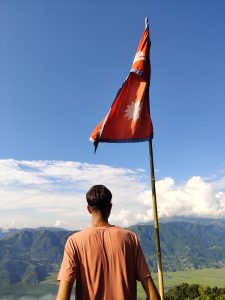 A person stands facing a mountainous landscape, holding a tall pole with the red flag of Nepal.