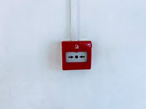A red square fire alarm call point is mounted on a plain white wall, with a white wire running vertically above it.