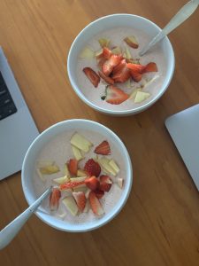 Two bowls of a creamy dish topped with slices of red strawberries and green apple are placed on a wooden table. 