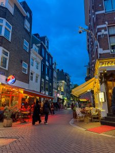 A lively European street scene at dusk in Amsterdam with people walking and sitting at outdoor café tables under warm lights. Tall, narrow buildings with large windows line the cobblestone street, and signs from nearby restaurants and bars glow against the deep blue evening sky.