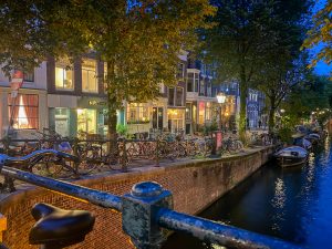 A charming evening canal scene with rows of bicycles parked along the brick edge, illuminated by glowing street lamps. Boats rest on the water, and behind them are tall, narrow buildings with lit windows and storefronts, framed by leafy trees under a twilight sky.