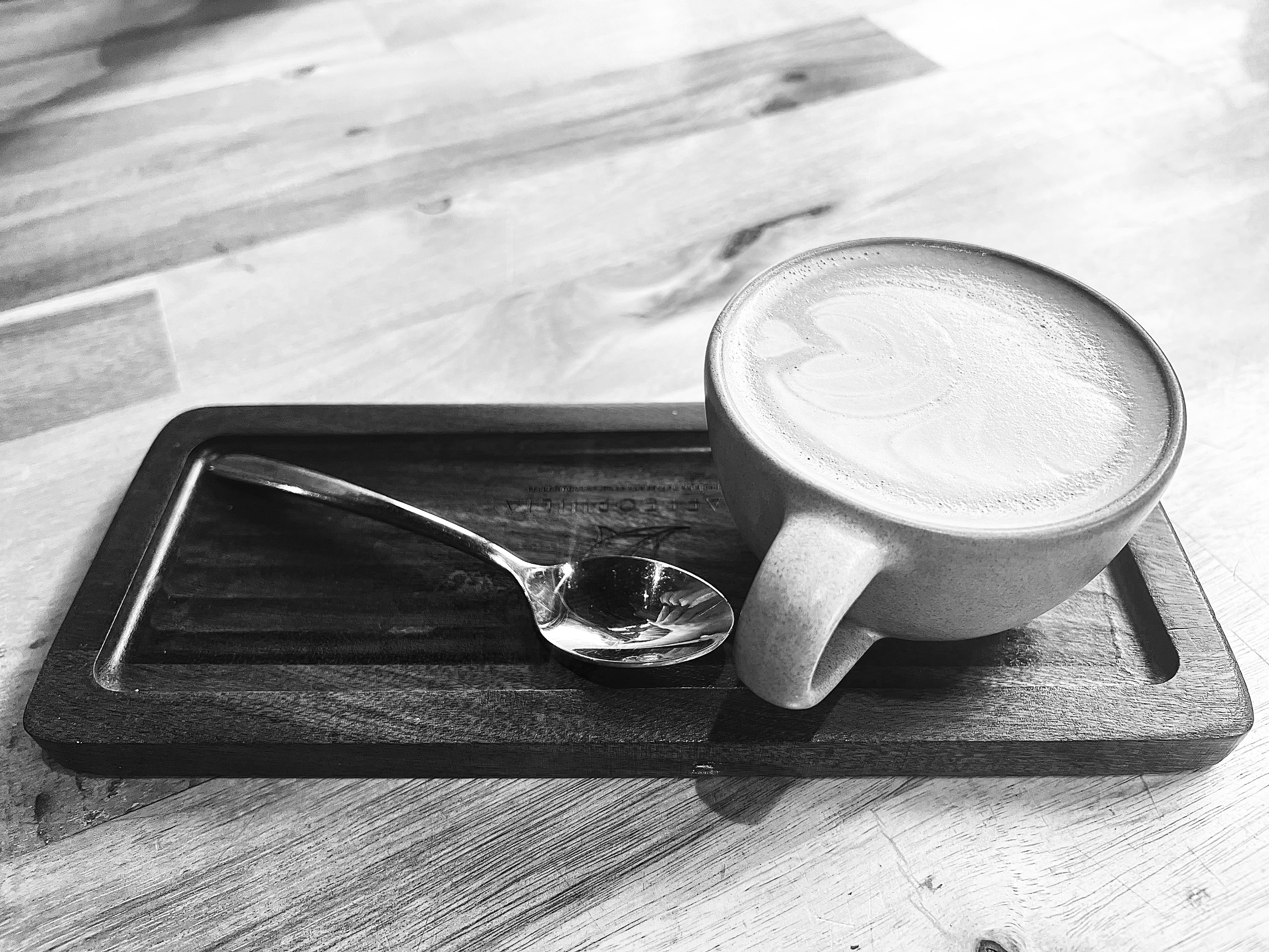 A black and white image of a cappuccino with latte art in a white cup on a wooden tray. A spoon lies beside, creating a calm, cozy ambiance.