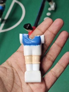 Hand holding the lower half of a broken foosball player figurine with a blue jersey, missing its upper body, against a green tabletop soccer field.
