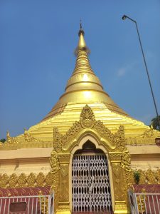 A golden pagoda in Lumbini, Nepal, with a conical spire, ornate decorative elements, and a closed gate in the foreground.