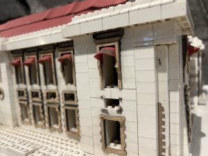 A close-up view of a detailed model building constructed with white bricks, featuring a red roof and decorative elements. The façade displays several windows with archways, some adorned with red awnings. The structure shows signs of wear, with exposed layers and texturing that adds a realistic appearance.