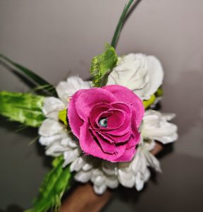 A close-up image of a colorful artificial flower arrangement featuring a prominent bright pink rose in the center, surrounded by white petals and green leaves. 