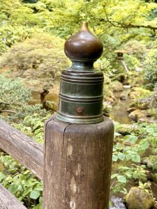 A close-up of a decorative bronze cap on a wooden post, set against a peaceful stream and green trees in the Japanese Garden, Portland. 