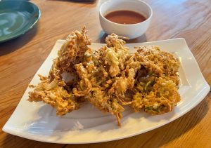 A serving of three golden-brown, crispy fried vegetable fritters -Pakoda, is presented on a white rectangular plate with a small bowl of red dipping sauce in the background.