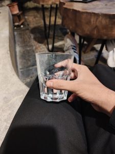 A hand holds a glass of water on black pants, with tables and floor in the background.