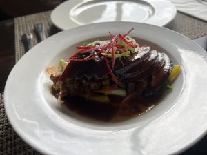 A close-up of a plate with a savory-looking pork belly dish topped with thinly sliced vegetables.
