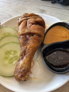 A close-up of a fried chicken drumstick placed on a white plate, accompanied by sliced cucumbers and two small bowls of dipping sauces.