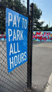 On the left, a large sign on a fence says "PAY TO PARK ALL HOURS". On the right, a parking lot with some graffiti in the background.