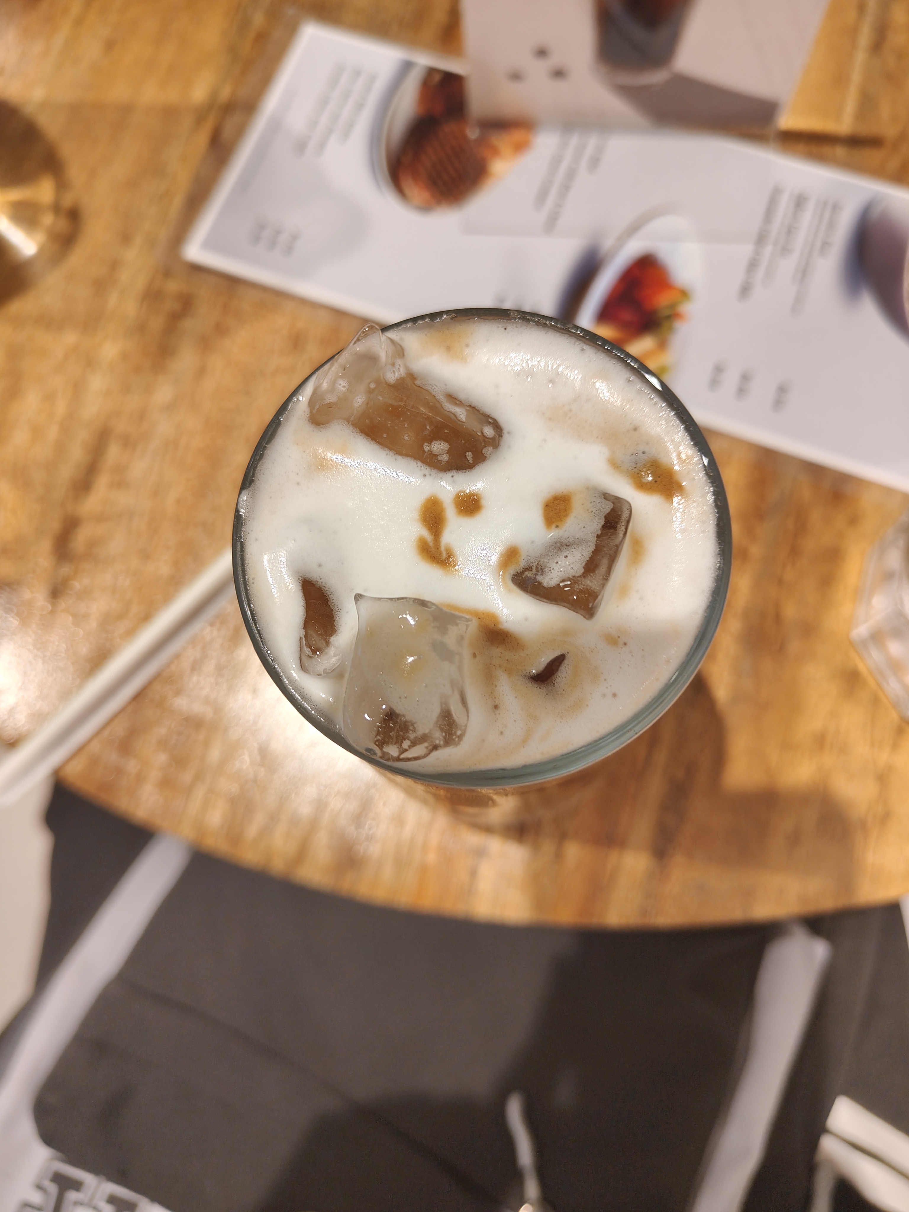 A glass of iced coffee with visible ice cubes and frothy milk on top sits on a wooden table