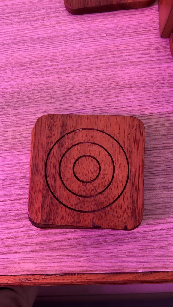 A wooden square coaster featuring three concentric circles etched into its surface.