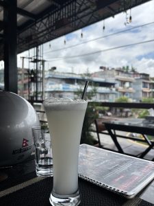 A tall glass of a cold white beverage with a straw sits on a table at an outdoor café. Next to it is a clear empty glass, a white motorcycle helmet with a Nepali flag logo, and a menu on the table. In the background, blurred buildings and greenery are visible under a partly cloudy sky.