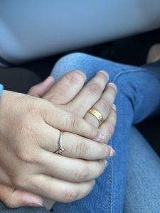 A close-up of two intertwined hands, one with a textured gold band and the other with a delicate stone-studded ring.
