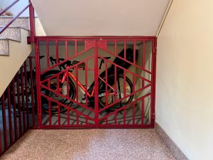 A red metal gate with a geometric pattern secures a small storage area under a staircase. Inside the enclosure, two bicycles are visible, one with a bright orange frame and the other primarily black, along with some other items partially obscured. The flooring is made of a light-colored material, and the wall is a muted shade. Steps leading to the upper level are seen to the left.