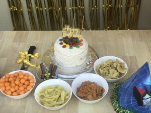 A small, white homemade birthday cake with "Happy Birthday" candles and various bowls of snacks, like chips and candy.
