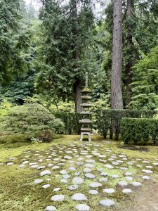 A stone pagoda stands in a circular pattern of moss and stone, surrounded by tall evergreens and lush shrubs within the tranquil Japanese Garden in Portland.