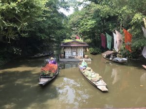 Several small, wooden boats on a calm river, with one boat carrying flowers and another carrying sacks. A small house is in the middle of the river.