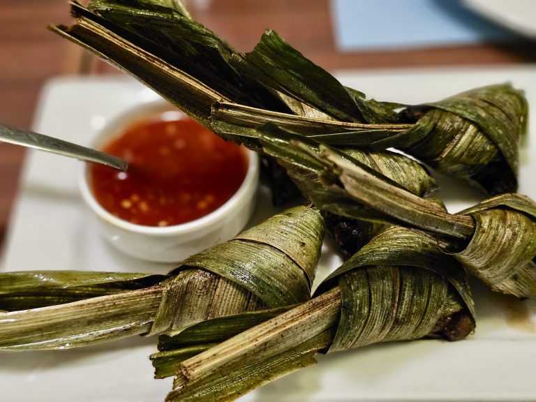 Fried chicken wrapped in pandan leaves, served with a bowl of sweet chili dipping sauce. The dish is placed on a white plate with intense indoor lighting.