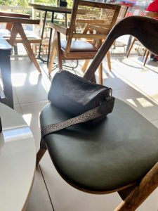 A dark leather handbag with a patterned strap rests on the olive-green padded seat of a wooden chair in a sunlit cafe.