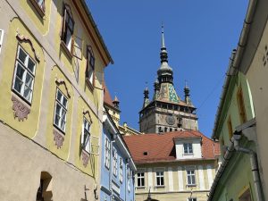 A view down a street of old town of Sighișoara (Romania) at a clock tower (Turnul cu Ceas) from Stradela Cet&abreve;ții. To the sides are buildings painted with yellow, blue, tan, and green. The buildings have terra cotta roof tiles.
