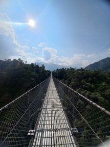 A long suspension bridge stretches into the distance, with the ground obscured below. The bridge features a metal walkway and is surrounded by lush green hills on either side. Above, the sky is bright and blue, with a few fluffy white clouds, and the sun shines brightly, casting rays of light across the scene.