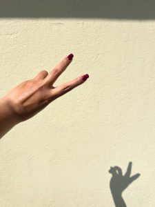 A hand with dark red nail polish is shown making a peace sign against a light-colored wall.