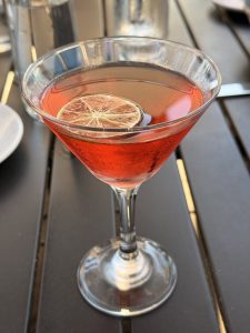 A close-up of a red cocktail served in a martini glass, topped with a dried lime slice. The drink sits on a black outdoor table with soft daylight reflections in the glass. 